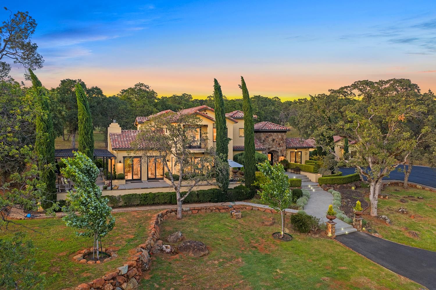 Wide twilight view of the Mediterranean estate from the lower lawn, the home's stucco facade and tile roof glowing softly through the oaks and cypress