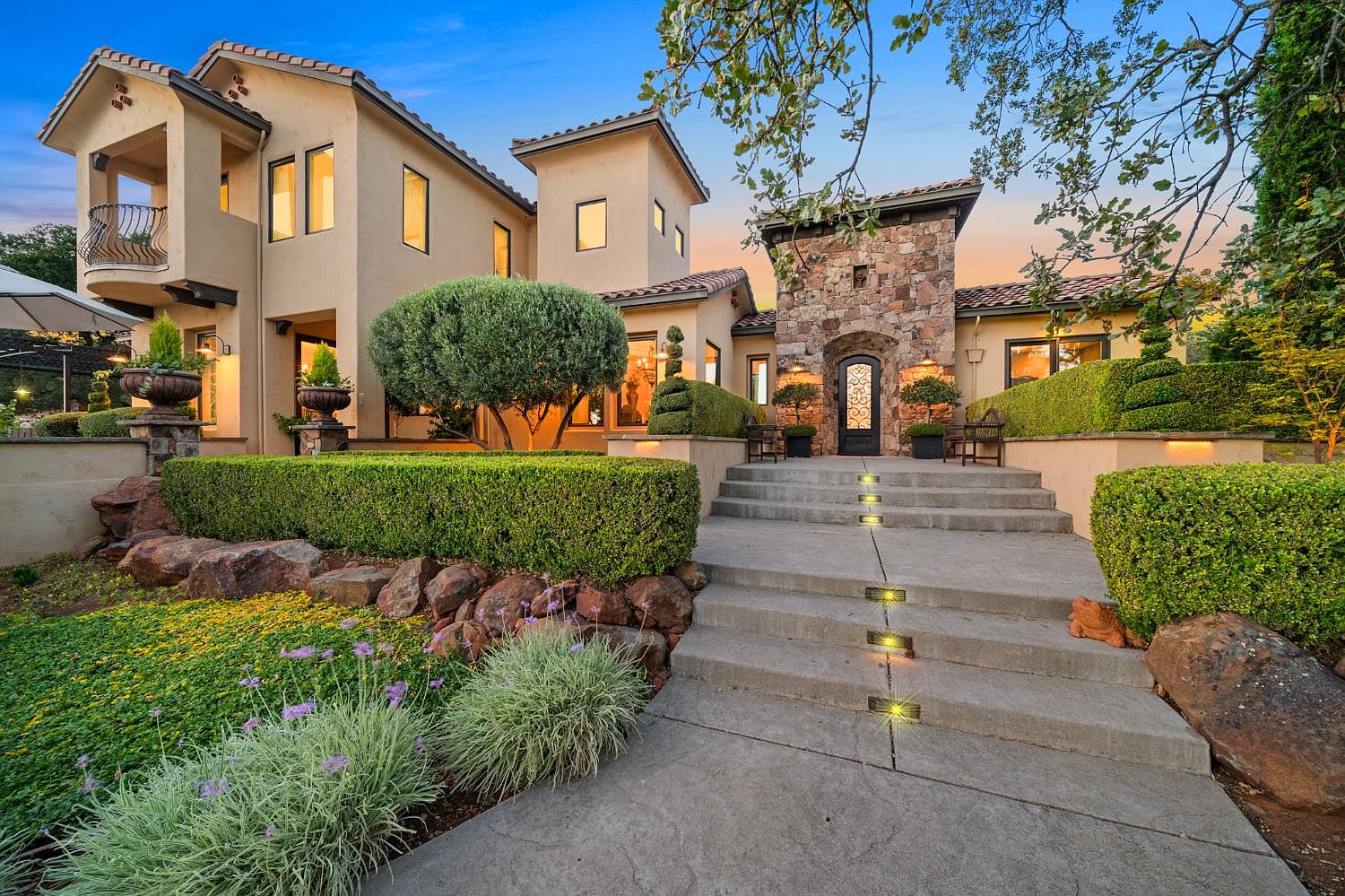 Front approach at twilight, illuminated stone steps leading to the carved wood front door framed by the home's stucco wings and topiary