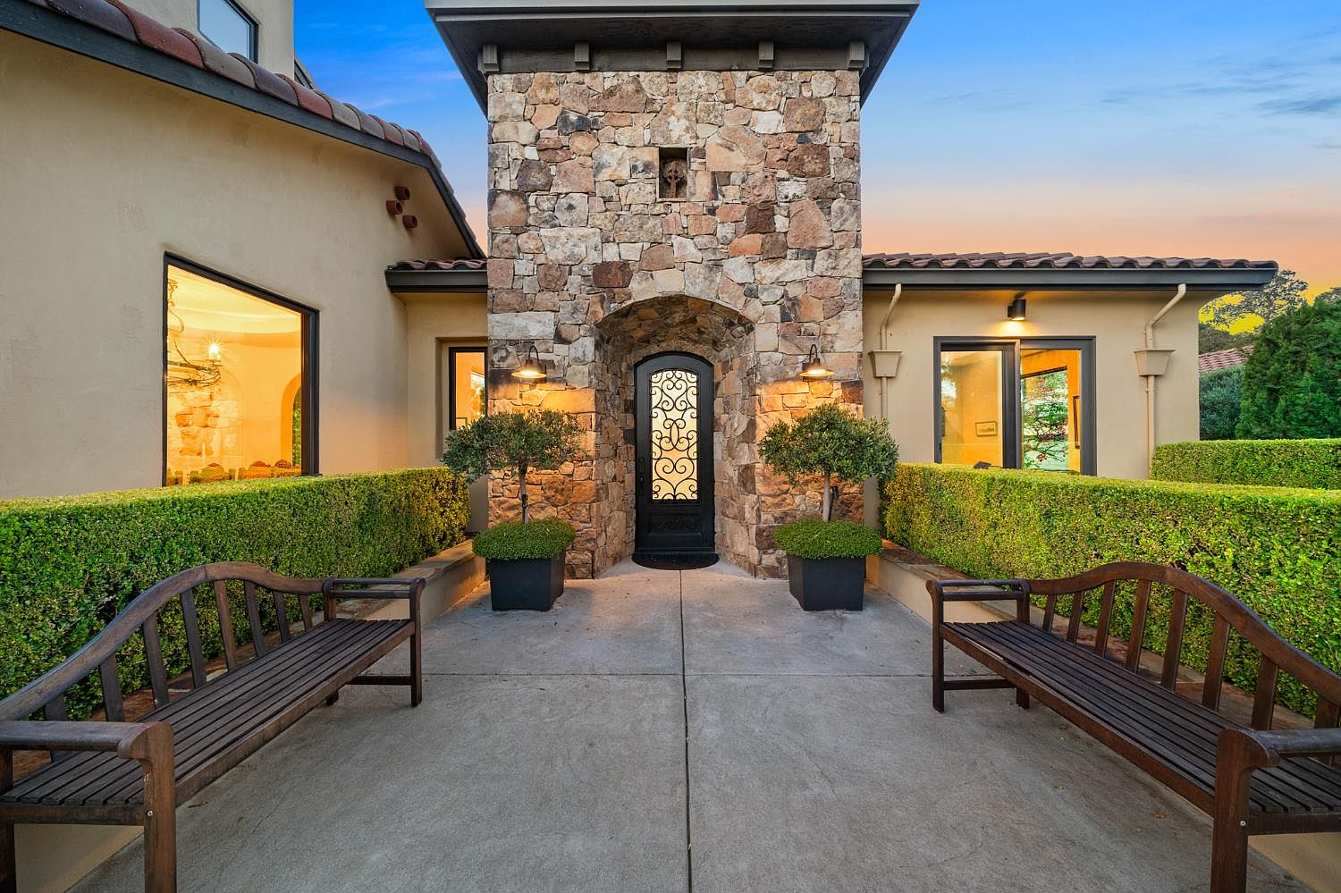 Detail of the stone entry tower with iron-grille door, paired potted boxwoods, sconces, and benches in the entry courtyard