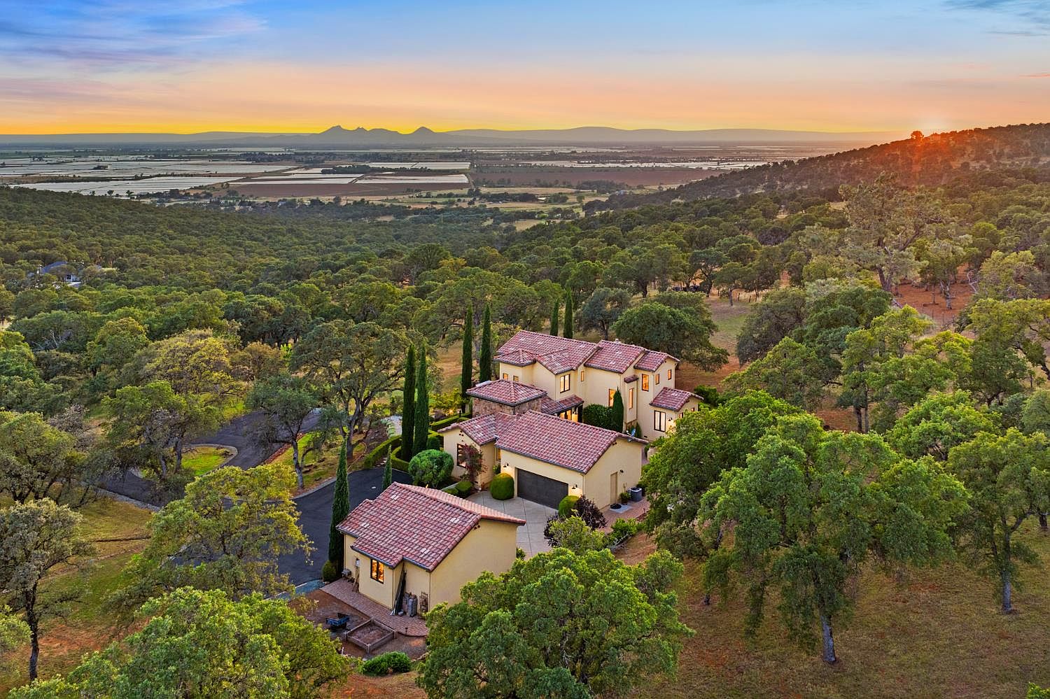 Aerial twilight view of 5133 Oak Grove Ct, a Mediterranean stucco-and-stone estate held above the Sacramento Valley with sunset light over distant mountains and a wide oak savanna foreground